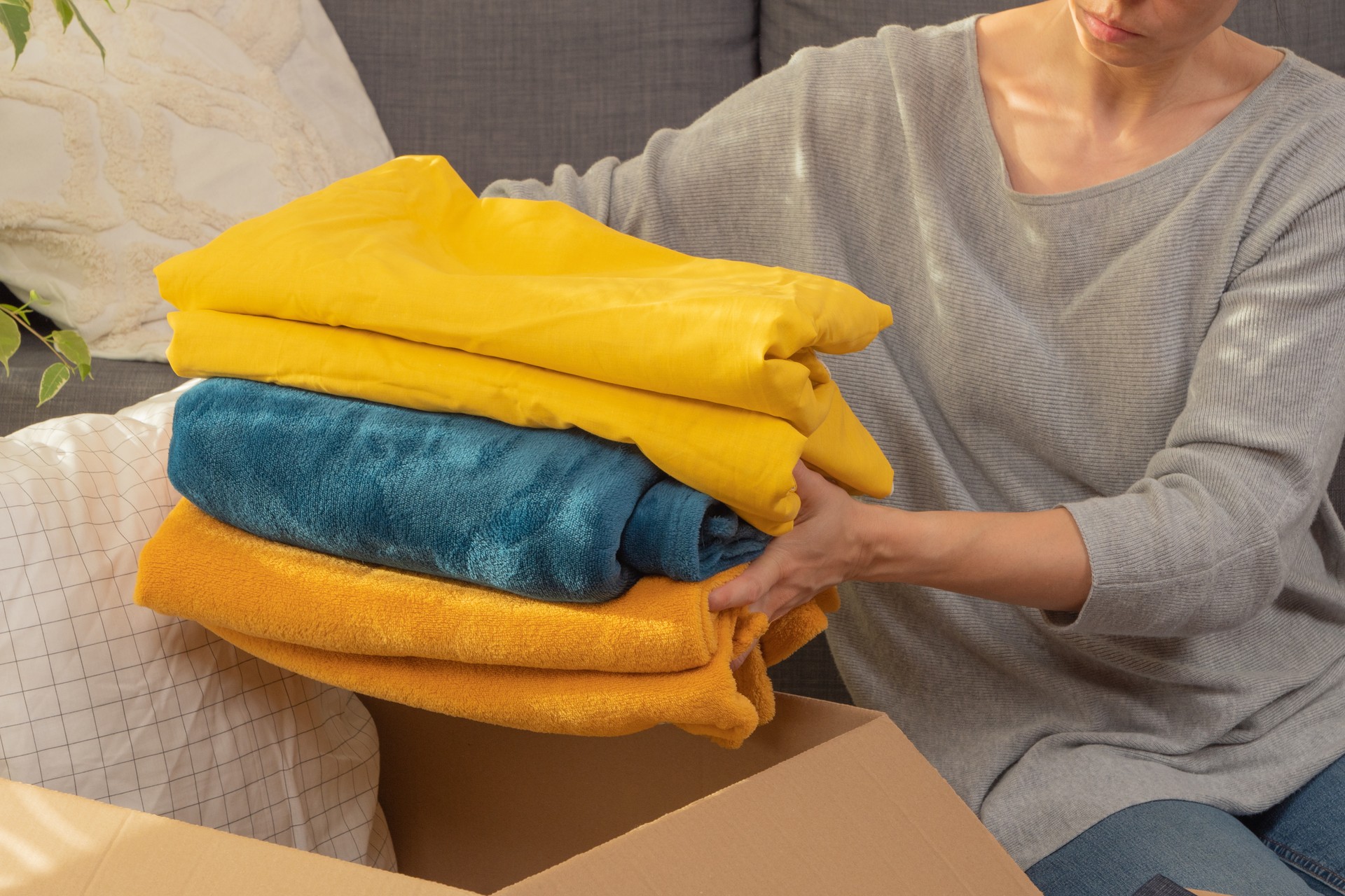 Female hands holding stack of bed linen. Woman preparing items and packing into cardboard box. Donations for charity, help low income families, declutter home, sell online, sustainable living concept