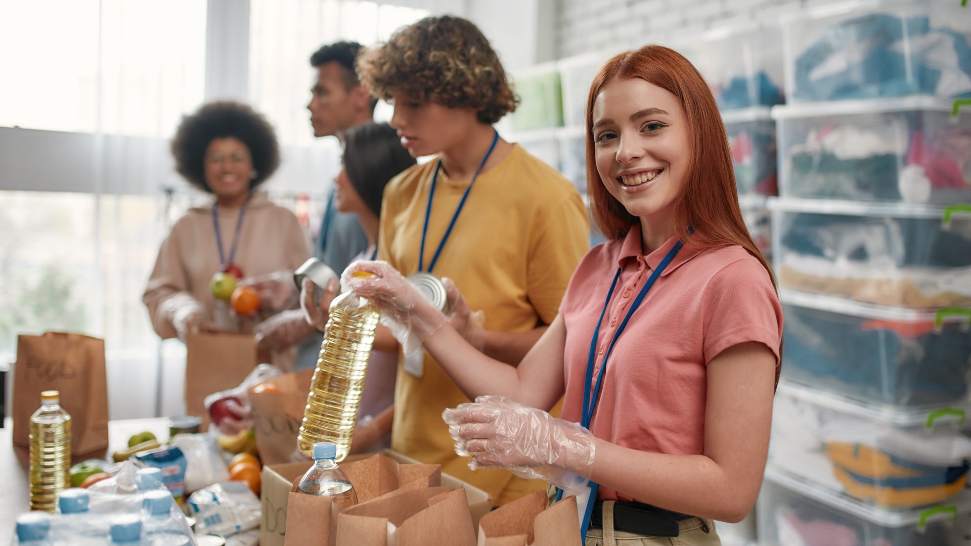 Young female volunteer smiling at camera while packing food and drinks donation into paper bags and box, Small group of people working in charitable foundation